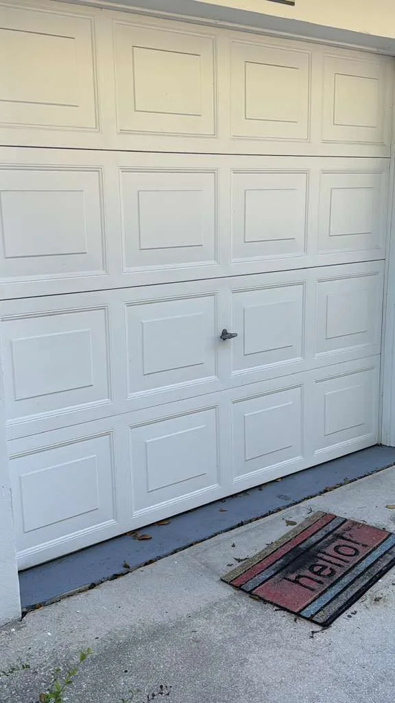 Residential white garage door jammed crooked with open gap along concrete foundation threshold